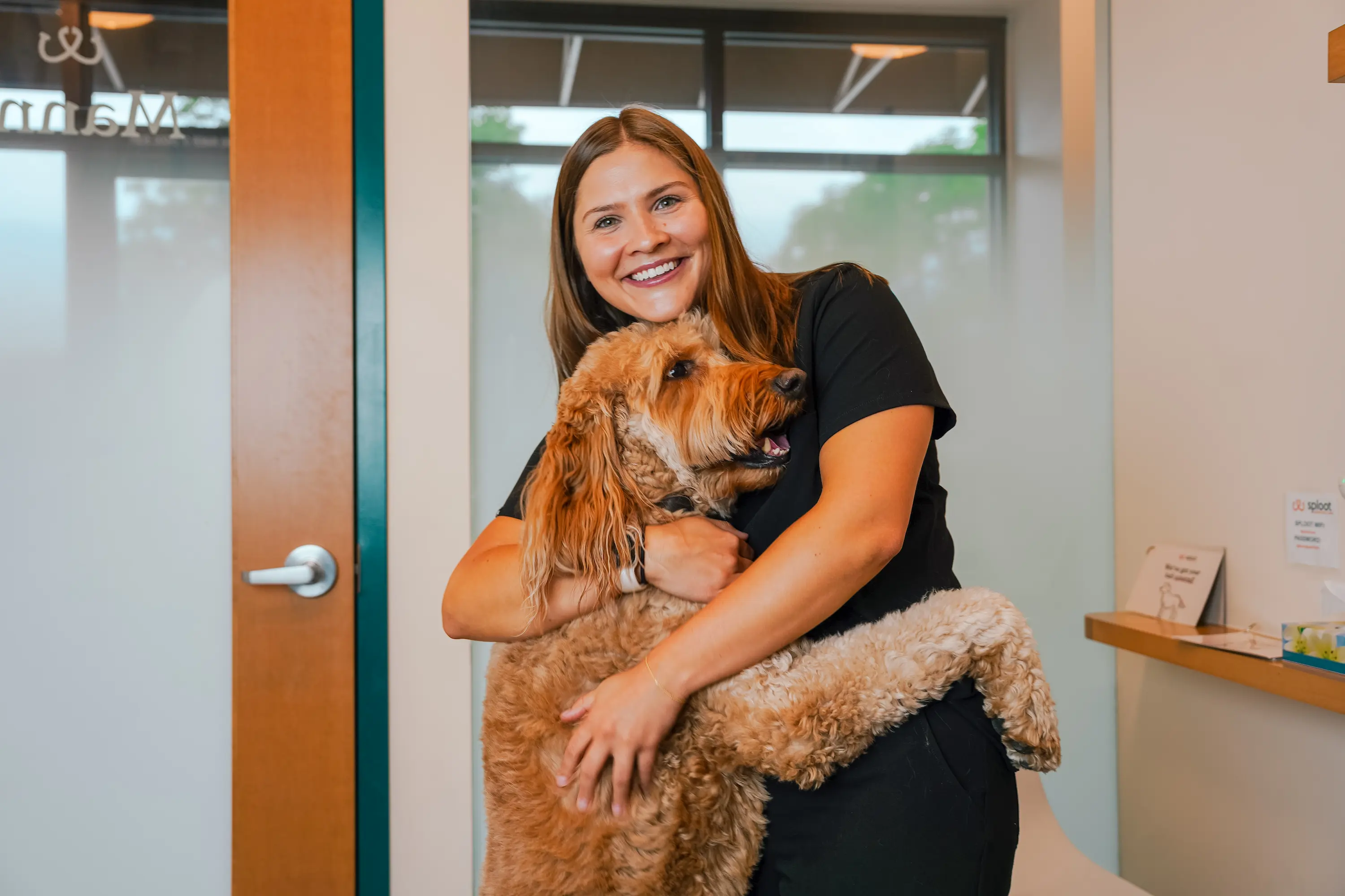 Smiling woman in a black outfit hugging a large curly-haired brown dog inside a Sploot clinic.